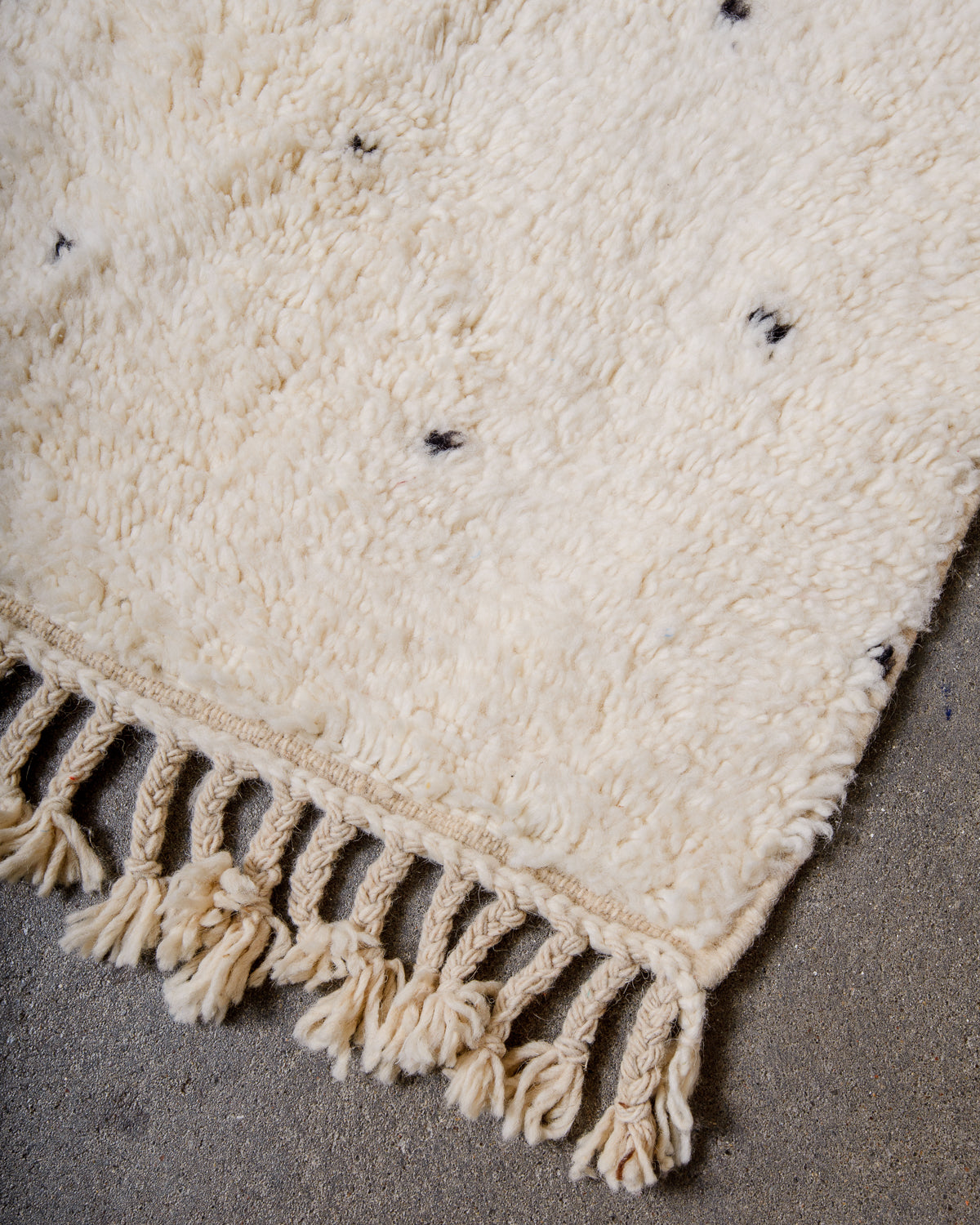 Corner of cream Beniourain wool rug with a tightly woven edge and natural wool tassels laying on concrete floor.